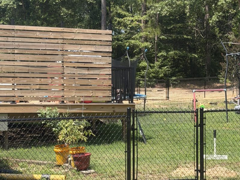 Black chain link fence with gate enclosing residential property during deck construction, showing natural wood materials and elevated structure on metal supports.