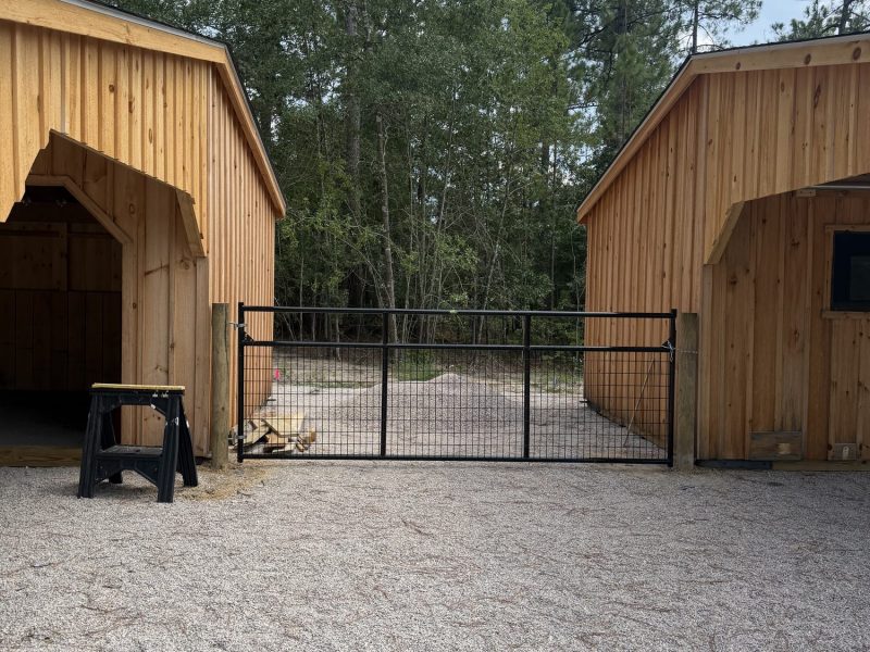 Black metal farm gate with mesh fencing installed between wooden barn structures and vinyl or wood post construction on rural property.