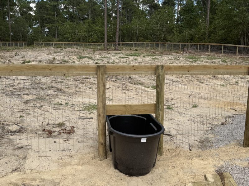 Black water trough in sandy equestrian arena surrounded by completed wood fencing installation at farm facility.