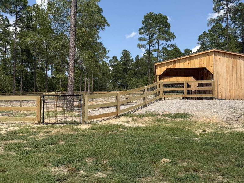 Horse barn with tan wood finish and open-sided design surrounded by wooden ranch-style fencing on rural property.