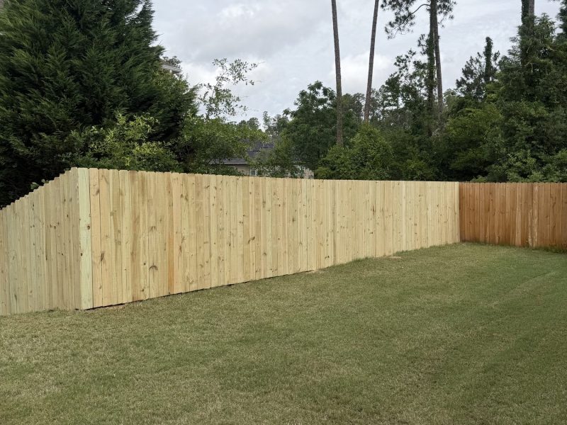 Newly installed wood privacy fence with vertical planks in residential backyard, lush landscaping and trees visible.