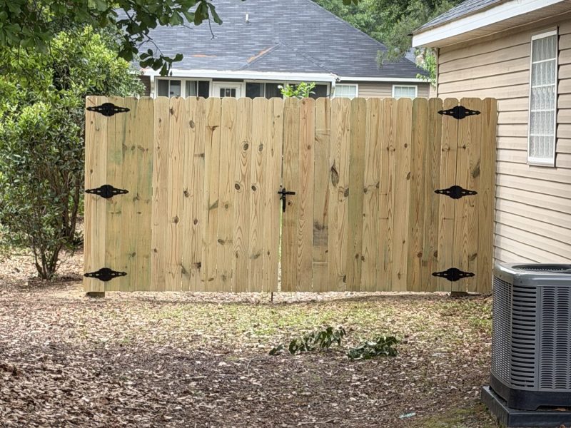 Light-colored wood privacy fence with vertical pickets and decorative black hinges separating residential backyards in Aiken area.