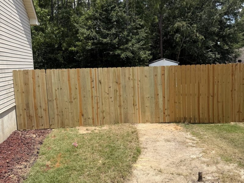 Light-colored wood privacy fence with vertical planks installed in residential backyard, separating property from wooded area.