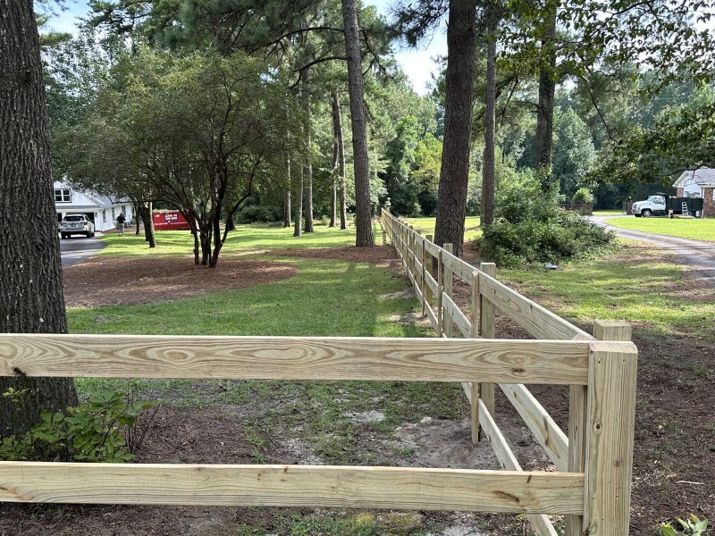 Newly installed treated wood rail fencing along rural property driveway with farm house and barn in background.