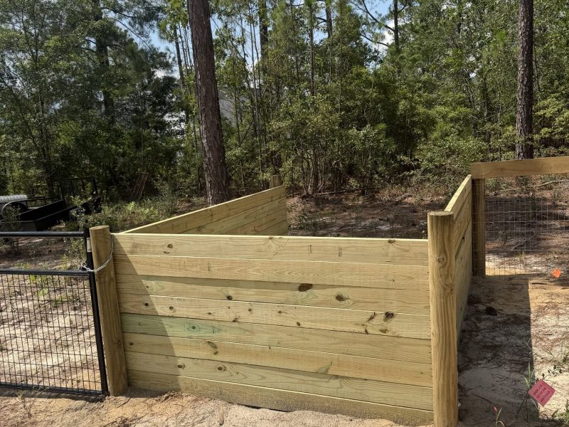 Pressure-treated wooden livestock feeding trough on rural property with chain link fencing installation in background.