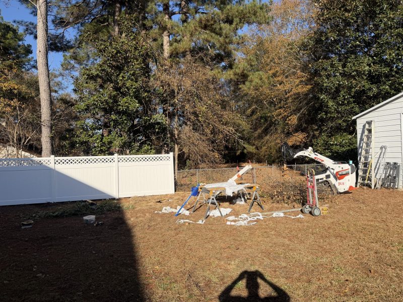 Residential backyard with newly installed white vinyl privacy fence, equipment, and shed during fence construction project.