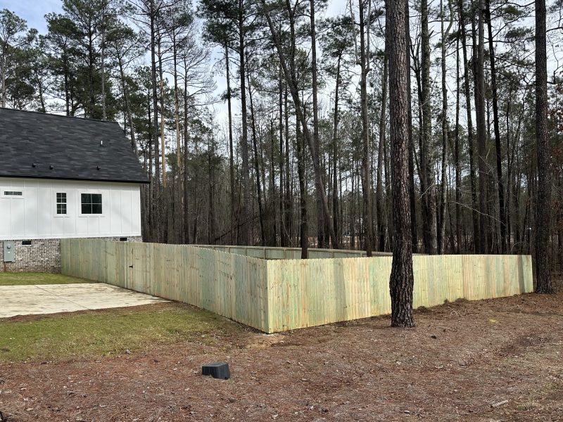 Residential wood fence installation in tan finish securing backyard perimeter beside white brick home with mature pines.