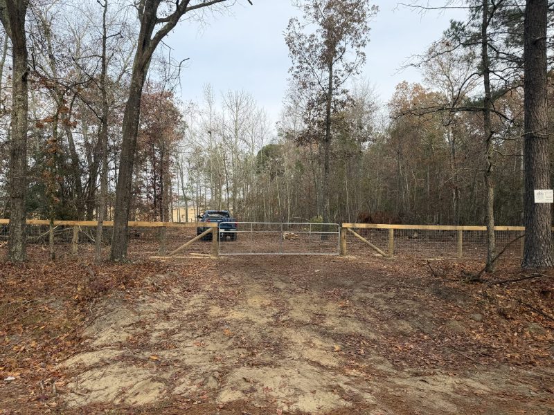 Wooden and metal swing gate entrance with tan privacy fence panels for rural property in wooded setting.