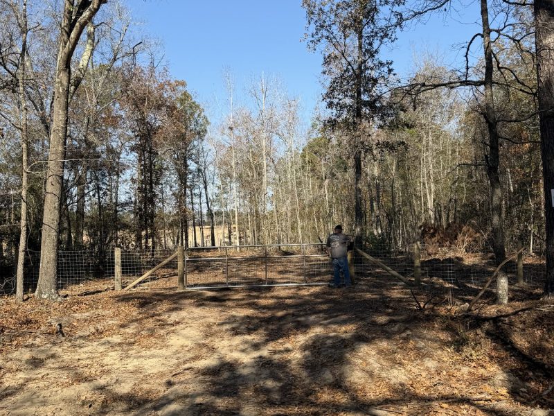 Wooden and wire farm fence lining a rural driveway entrance with metal gate in Aiken County.