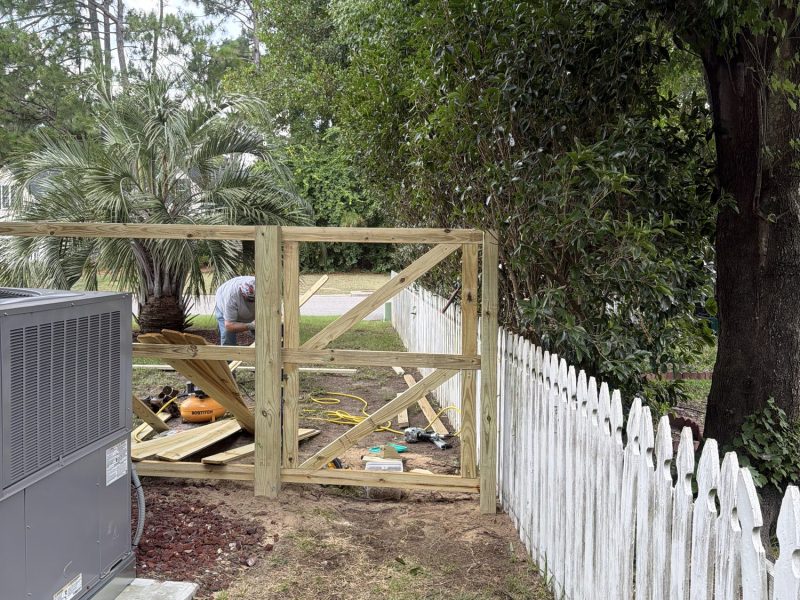 Wooden gate with X-braced panels under construction next to white picket fence in residential side yard.